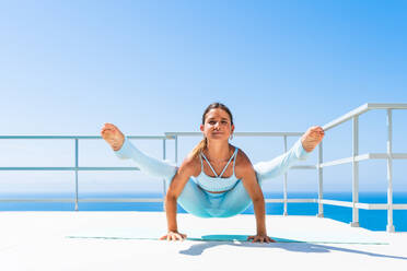 Beautiful young atlethic woman wearing sportswear doing yoga meditation on the terrace rooftop with sea view and blue sky - Yoga practicer training outdoors, healthy lifestyle, sport and meditation concept - DMDF03754