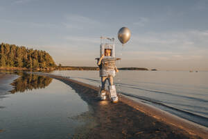 Boy in astronaut costume holding silver balloon at beach - EVKF00022