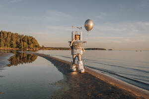 Junge im Astronautenkostüm hält silbernen Luftballon am Strand - EVKF00022