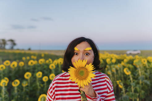 Reife Frau mit Blütenblättern auf den Augenbrauen, die den Mund mit einer Sonnenblume bedecken - IEF00485