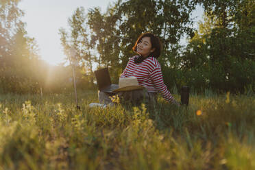 Smiling mature woman with hat sitting on grass in field - IEF00500