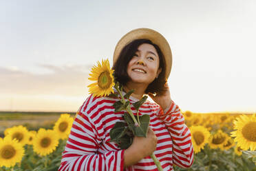 Smiling woman with sunflower wearing hat in field - IEF00511