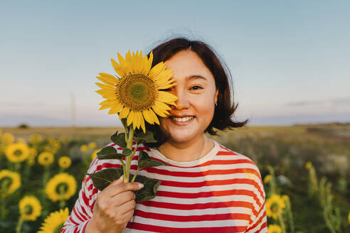 Lächelnde Frau bedeckt Auge mit Sonnenblume bei Sonnenuntergang - IEF00515