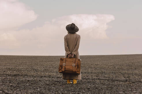 Frau mit braunem Koffer in gepflügtem Feld stehend - VSNF01370
