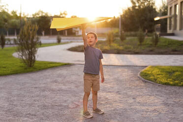 Boy holding paper airplane and standing in park - ONAF00632