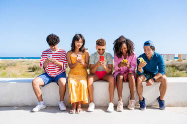 Multiethnic group of young happy friends bonding outside, having fun on summertime vacation - Multicultural cheerful people with summer clothes using mobile phones, concepts about youth, friendship and communication technology - DMDF03897