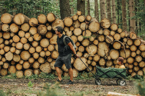 Father pulling son in cart by wood logs in forest - VSNF01383