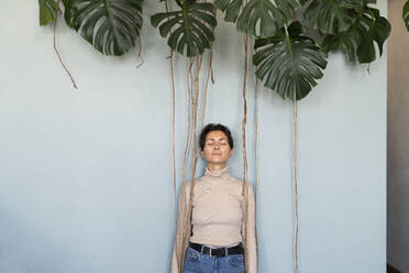 Smiling woman with eyes closed leaning on wall under monstera plant - VPIF08553