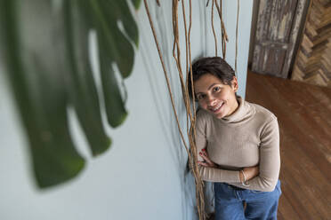 Happy woman with arms crossed leaning on wall by vines - VPIF08556