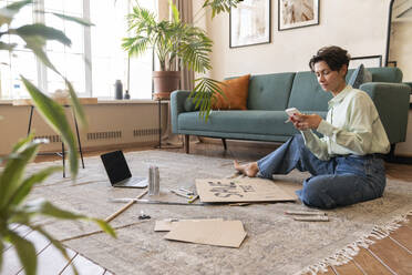 Activist making environment placard and using smart phone on carpet in living room at home - VPIF08578