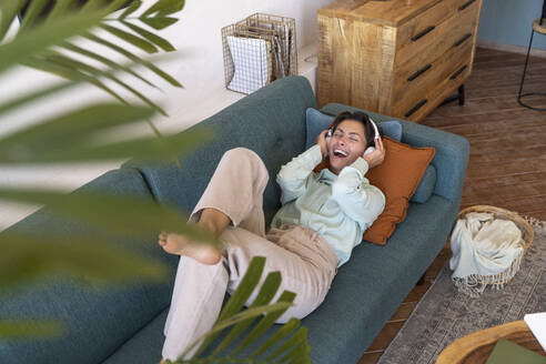 Happy woman wearing wireless headphones listening to music on sofa - VPIF08600