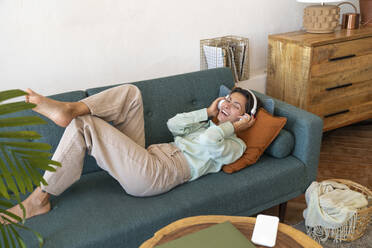 Smiling woman wearing wireless headphones enjoying music on sofa - VPIF08601
