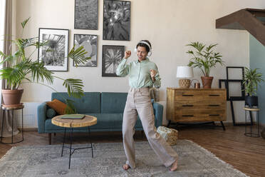 Happy woman wearing wireless headphones dancing in living room at home - VPIF08606