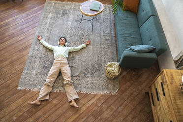 Thoughtful woman wearing wireless headphones lying on carpet at home - VPIF08608