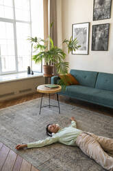 Happy woman wearing wireless headphones relaxing on carpet at home - VPIF08610