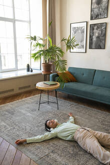 Glückliche Frau mit drahtlosen Kopfhörern, die sich zu Hause auf dem Teppich entspannt - VPIF08610