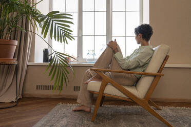 Thoughtful woman sitting in armchair near window at home - VPIF08625