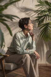 Thoughtful woman sitting in armchair amidst plants - VPIF08636