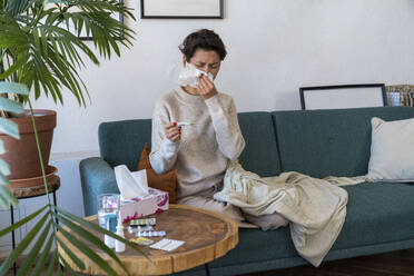 Sick woman blowing nose and sitting on sofa at home - VPIF08638