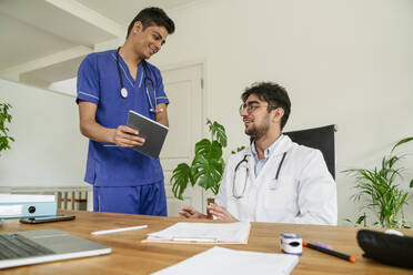 Ein lächelnder Arzt zeigt einem Kollegen am Schreibtisch in einer Klinik einen Tablet-PC - OSF02078