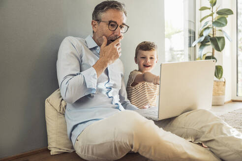 Mann mit Hand auf dem Mund bei der Benutzung eines Laptops durch seinen Enkel zu Hause - UUF29981