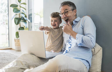 Cheerful boy pointing at laptop sitting by grandfather in living room - UUF29982