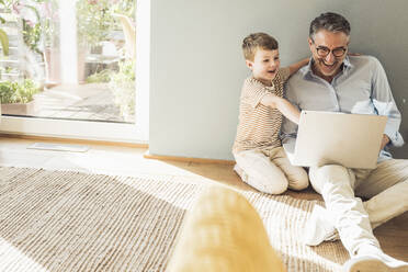 Smiling grandfather using laptop with grandson in living room - UUF29984