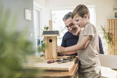 Smiling man making wooden birdhouse with grandson at home - UUF29987