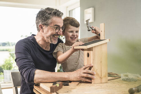 Cheerful boy timbering birdhouse by grandfather at home - UUF29997