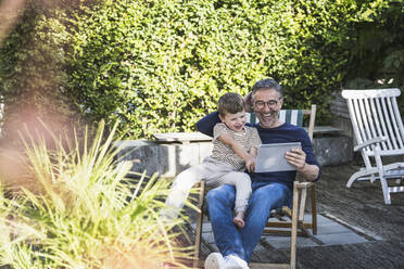 Happy man and boy using tablet PC sitting at back yard - UUF30008