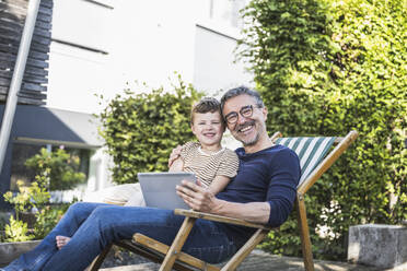 Smiling grandfather and grandson sitting with tablet computer in back yard - UUF30010