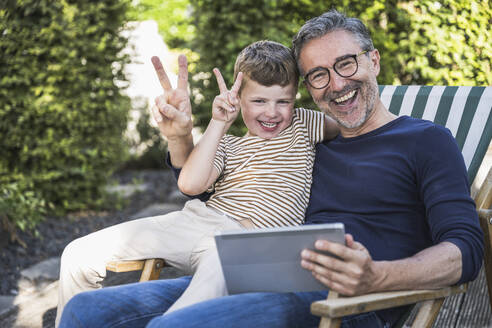 Cheerful man and boy gesturing peace sign sitting with tablet PC - UUF30012