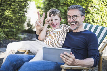 Glücklicher Mann und Junge mit Friedenszeichen sitzend mit Tablet-PC - UUF30013