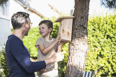 Junge befestigt Vogelhaus auf Baum und spricht mit Großvater im Hinterhof - UUF30014