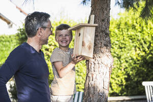 Happy boy fixing birdhouse on tree by grandfather in back yard - UUF30019