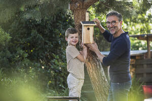 Smiling man and boy hanging birdhouse on tree in back yard - UUF30022