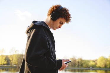 Jugendlicher, der im Park ein Mobiltelefon benutzt und über Kopfhörer Musik hört - EYAF02742