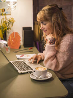 Smiling freelancer wearing sweater using laptop at home - LAF02825