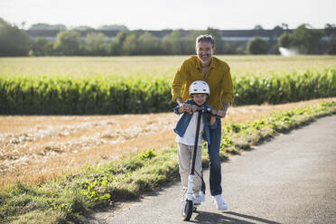 Smiling grandson riding push scooter with grandfather on road - UUF30045
