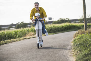 Happy man enjoying riding push scooter with grandson on street - UUF30046