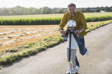 Happy grandson riding push scooter with grandfather on road - UUF30047