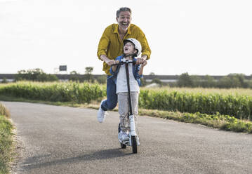 Cheerful grandfather enjoying riding push scooter with grandson on street - UUF30048