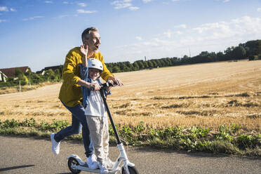 Cheerful man enjoying riding push scooter with grandson on sunny day - UUF30051