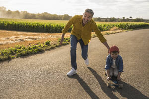 Happy man pushing grandson on skateboard on road - UUF30057