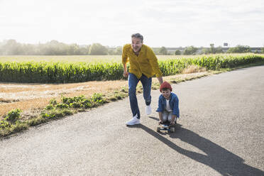 Glücklicher Großvater schiebt seinen Enkel auf dem Skateboard über die Straße - UUF30058