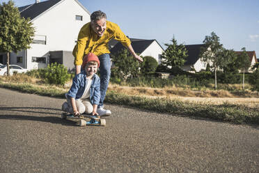 Cheerful man pushing grandson on skateboard at sunny day - UUF30059