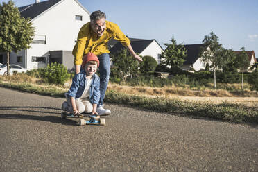 Fröhlicher Mann schiebt seinen Enkel auf dem Skateboard an einem sonnigen Tag - UUF30059