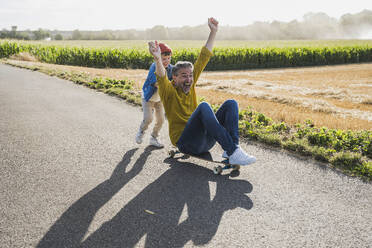 Happy boy pushing grandfather on skateboard on road - UUF30060