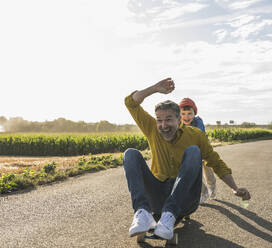 Cheerful grandson pushing grandfather on skateboard on road - UUF30061