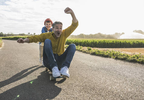 Boy pushing cheerful grandfather on skateboard on road - UUF30062